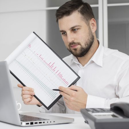 Man in a white shirt reviews a bound chart with pink and blue bars, pointing with a pen at a laptop on a desk.